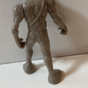 May include: A grey, textured figurine of a humanoid creature, viewed from the back. The figure has a rough, organic appearance with detailed musculature and a strap across the back. The feet are large and flat.