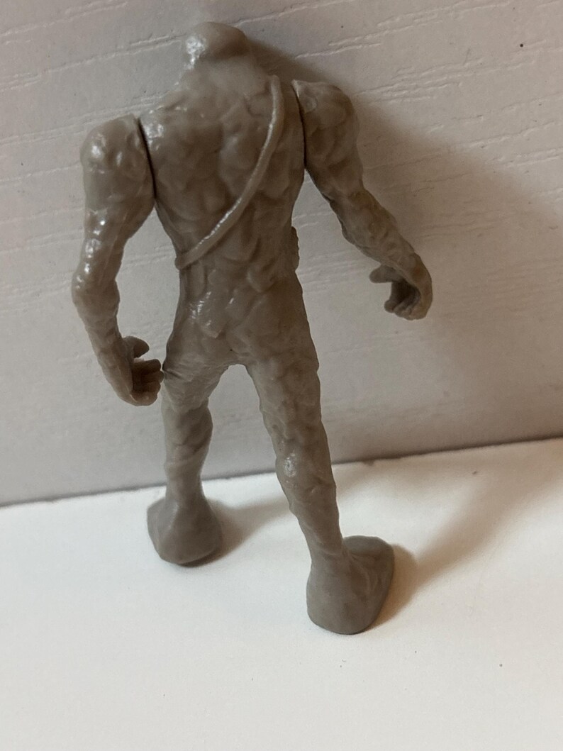 May include: A grey, textured figurine of a humanoid creature, viewed from the back. The figure has a rough, organic appearance with detailed musculature and a strap across the back. The feet are large and flat.