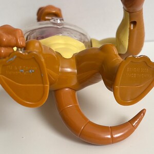 May include: A brown and yellow toy figure with a clear plastic head. The figure has a tail and is lying on its back. The feet of the figure are visible, and the words "BANDAI 2006 MADE IN CHINA" are printed on one foot.