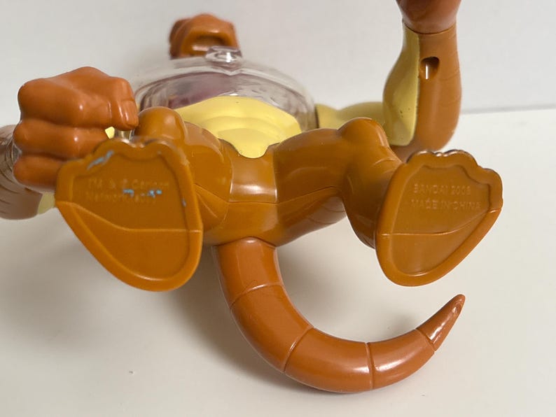 May include: A brown and yellow toy figure with a clear plastic head. The figure has a tail and is lying on its back. The feet of the figure are visible, and the words "BANDAI 2006 MADE IN CHINA" are printed on one foot.
