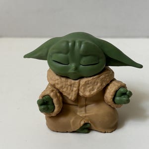 May include: A green figurine of a character with large ears, closed eyes, and small hands in a meditative pose. The figure wears a tan coat with a furry collar. The figurine is a collectible item.