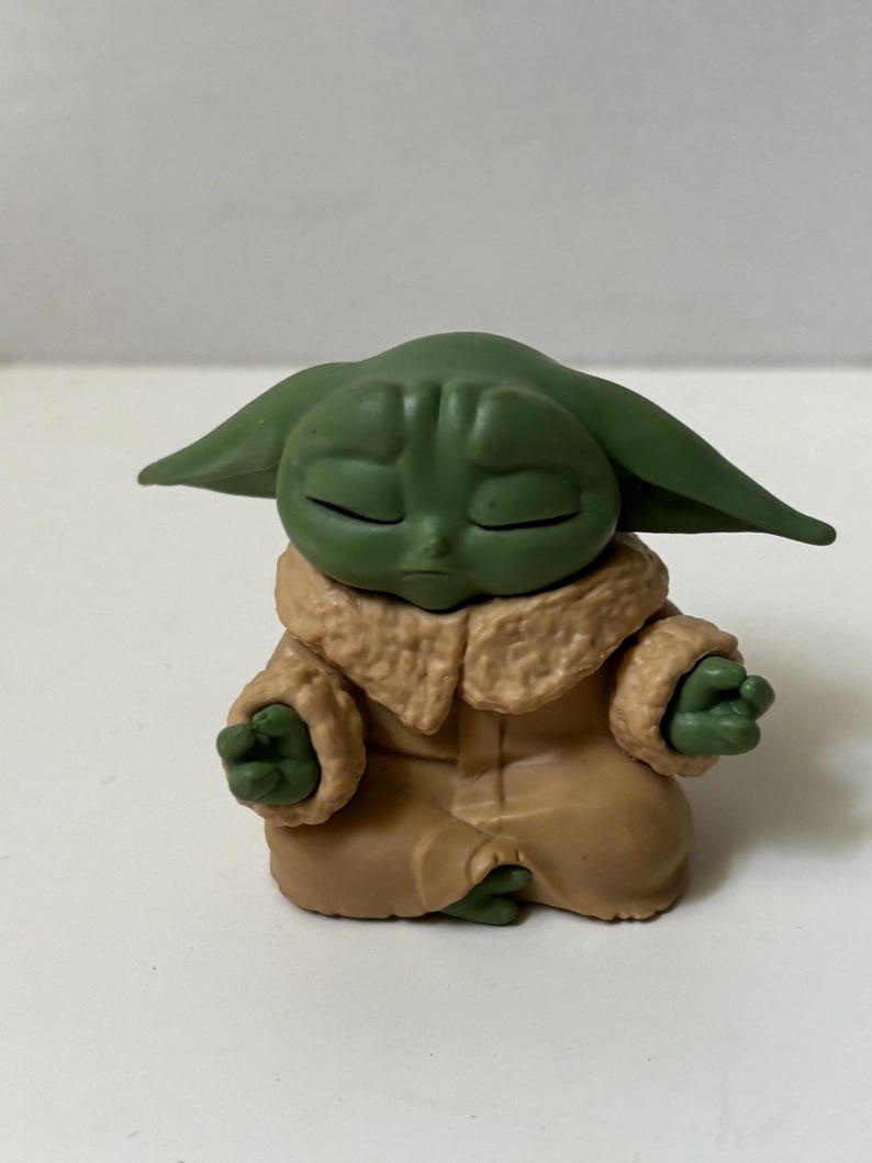May include: A green figurine of a character with large ears, closed eyes, and small hands in a meditative pose. The figure wears a tan coat with a furry collar. The figurine is a collectible item.
