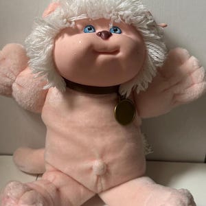 May include: A plush doll with light pink skin, bright blue eyes, and a small brown nose. The doll has a thick mane of white yarn hair, a dark brown collar with a round gold-colored metal tag, and a small belly button detail on its torso.