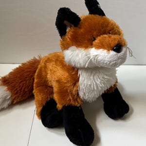 May include: A plush fox toy with orange, white, and black fur. The fox has a white chest and muzzle, black paws and ears, and a fluffy tail with a white tip. The toy has a realistic design and is suitable for children.