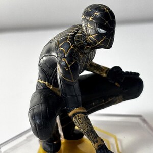Puede incluir: Una figura de Spider-Man negra y dorada en posici&oacute;n agachada. La figura tiene un dise&ntilde;o detallado con detalles dorados en el traje y un dise&ntilde;o de ojos blancos. La figura est&aacute; sobre una base transparente con detalles dorados.