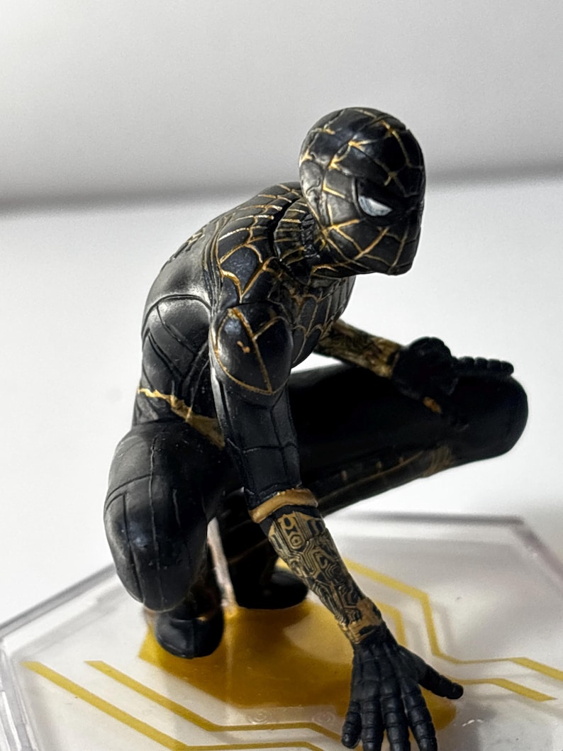 Puede incluir: Una figura de Spider-Man negra y dorada en posici&oacute;n agachada. La figura tiene un dise&ntilde;o detallado con detalles dorados en el traje y un dise&ntilde;o de ojos blancos. La figura est&aacute; sobre una base transparente con detalles dorados.