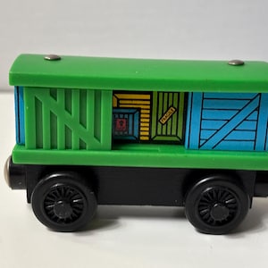 May include: A wooden toy train cargo car featuring a bright green roof and base, black wheels, and blue side panels with diagonal bracing. The center section displays a graphic of stacked yellow and green crates with the word FRAGILE printed on one.