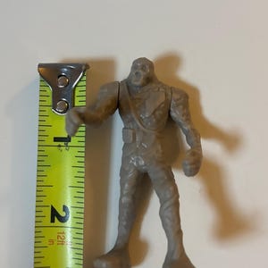 May include: A gray plastic monster action figure, approximately 6.35 cm tall, with a textured surface. The figure has a skeletal head and is posed with one arm extended. A yellow and black measuring tape is next to the figure.