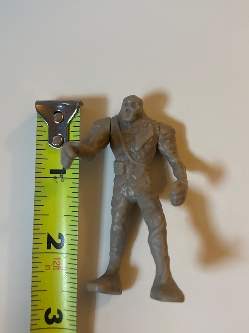 May include: A gray plastic monster action figure, approximately 6.35 cm tall, with a textured surface. The figure has a skeletal head and is posed with one arm extended. A yellow and black measuring tape is next to the figure.
