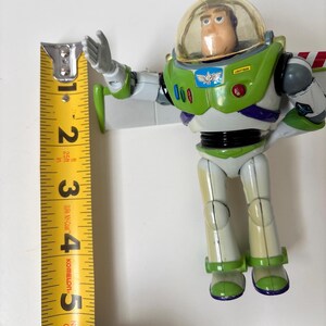 以下が含まれることがあります： 緑、白、紫を基調としたバズ・ライトイヤーのアクションフィギュア。透明なヘルメット付き。黄色い巻尺で測ると、フィギュアの高さは約13cm。ライトアップ機能付き。