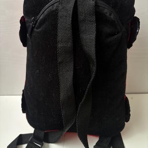 May include: A black and red backpack featuring a textured black fabric back panel, a top carry handle, and two adjustable black shoulder straps. The design includes a zippered main compartment and small side pockets with black flaps on each side.