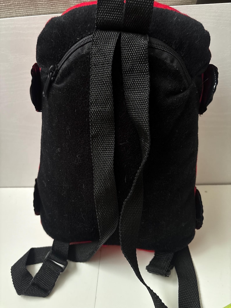 May include: A black and red backpack featuring a textured black fabric back panel, a top carry handle, and two adjustable black shoulder straps. The design includes a zippered main compartment and small side pockets with black flaps on each side.