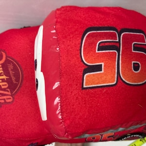 May include: A plush red toy car shaped like a race car with the number 95 printed in black and orange on the side. The car features a white windscreen, a Rust-eze logo on the bonnet, and a yellow measuring tape visible at the bottom right corner.