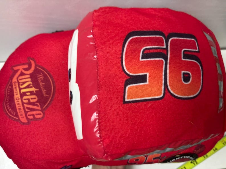 May include: A plush red toy car shaped like a race car with the number 95 printed in black and orange on the side. The car features a white windscreen, a Rust-eze logo on the bonnet, and a yellow measuring tape visible at the bottom right corner.