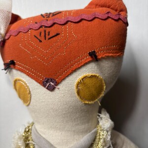 Puede incluir: Mu&ntilde;eca hecha a mano con una cabeza naranja con forma de zorro, con detalles bordados y un ribete rosa. La mu&ntilde;eca tiene c&iacute;rculos amarillos en las mejillas y un cuerpo color crema. Est&aacute; adornada con un cuello decorativo.