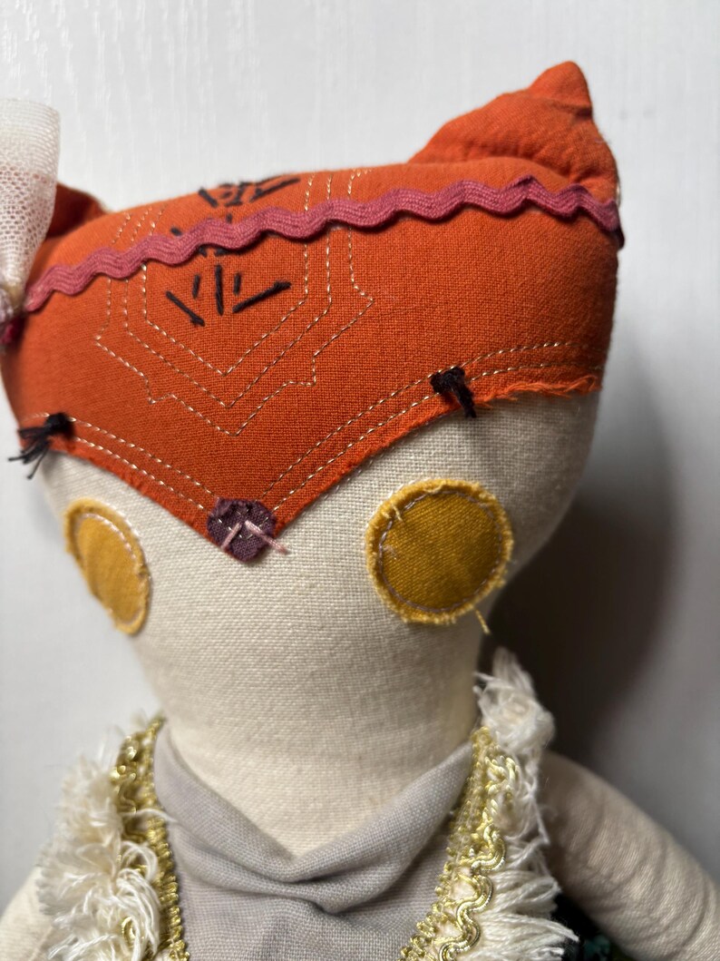 Puede incluir: Mu&ntilde;eca hecha a mano con una cabeza naranja con forma de zorro, con detalles bordados y un ribete rosa. La mu&ntilde;eca tiene c&iacute;rculos amarillos en las mejillas y un cuerpo color crema. Est&aacute; adornada con un cuello decorativo.