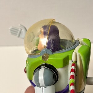以下が含まれることがあります： 透明なヘルメットと緑と白の宇宙服を着た、バズ・ライトイヤーのアクションフィギュア。フィギュアは紫色の頭と赤と白のストライプのアクセサリーを持っています。コレクション用の玩具です。