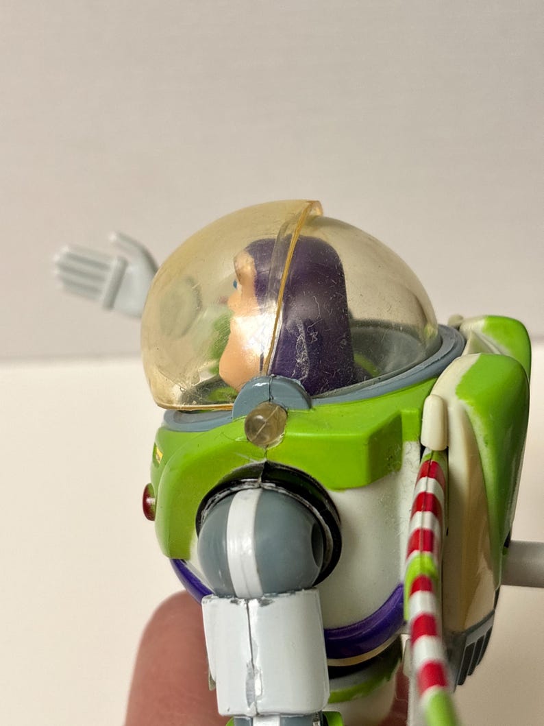 以下が含まれることがあります： 透明なヘルメットと緑と白の宇宙服を着た、バズ・ライトイヤーのアクションフィギュア。フィギュアは紫色の頭と赤と白のストライプのアクセサリーを持っています。コレクション用の玩具です。