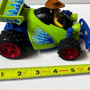 以下が含まれることがあります： 黒いタイヤと赤いリムが付いた、緑と青のおもちゃのレーシングカー。 トイ・ストーリーのウッディのフィギュアが運転席にいます。 車の長さは約13cmです。