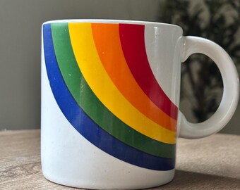 May include: White ceramic mug with a curved handle. The mug features a rainbow design with bands of blue, green, yellow, orange, and red. The mug is sitting on a wooden surface.