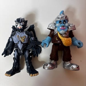 May include: Two action figures are displayed. One is a dark gray and black creature with wings, and the other is a blue-skinned figure in armor. The figures are on a white surface. The figures have detailed features and are likely toys.