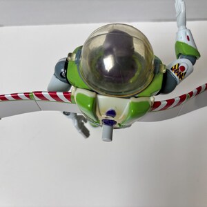 以下が含まれることがあります： 透明なヘルメットと腕を伸ばした緑と白のバズ・ライトイヤーのアクションフィギュア。翼は白地に赤と白のストライプ。フィギュアはトイ・ストーリーシリーズのおもちゃです。