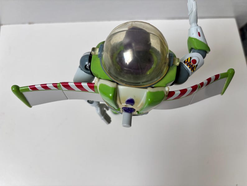 以下が含まれることがあります： 透明なヘルメットと腕を伸ばした緑と白のバズ・ライトイヤーのアクションフィギュア。翼は白地に赤と白のストライプ。フィギュアはトイ・ストーリーシリーズのおもちゃです。