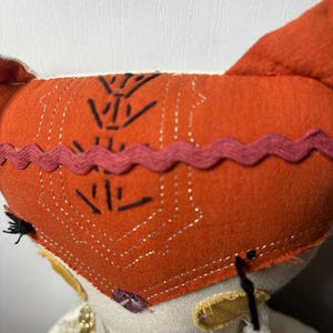 Puede incluir: Primer plano de una mu&ntilde;eca hecha a mano con un gorro naranja con dise&ntilde;os geom&eacute;tricos negros y un ribete rosa. La mu&ntilde;eca tiene una cara color crema, orejas amarillas y un cuello decorativo. Los ojos de la mu&ntilde;eca son negros.