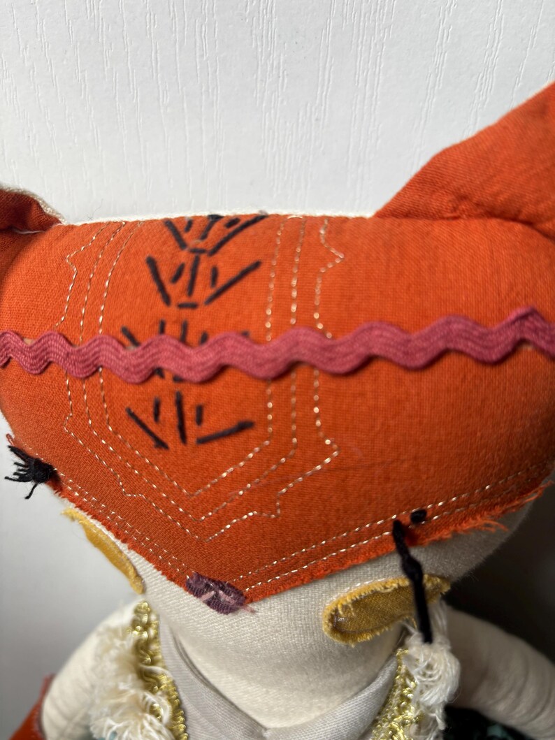 Puede incluir: Primer plano de una mu&ntilde;eca hecha a mano con un gorro naranja con dise&ntilde;os geom&eacute;tricos negros y un ribete rosa. La mu&ntilde;eca tiene una cara color crema, orejas amarillas y un cuello decorativo. Los ojos de la mu&ntilde;eca son negros.