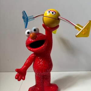 May include: A red Elmo figurine holding a yellow and white toy with blue and yellow accents. The Elmo figure has large white eyes, an orange nose, and an open mouth. The toy has a smiley face.