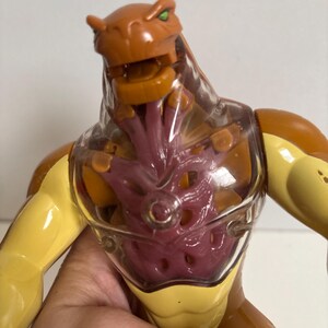 May include: A brown and yellow action figure with a clear chest cavity revealing a purple interior. The figure has a reptilian head with green eyes and is posed with its arms extended. The toy is made of plastic.