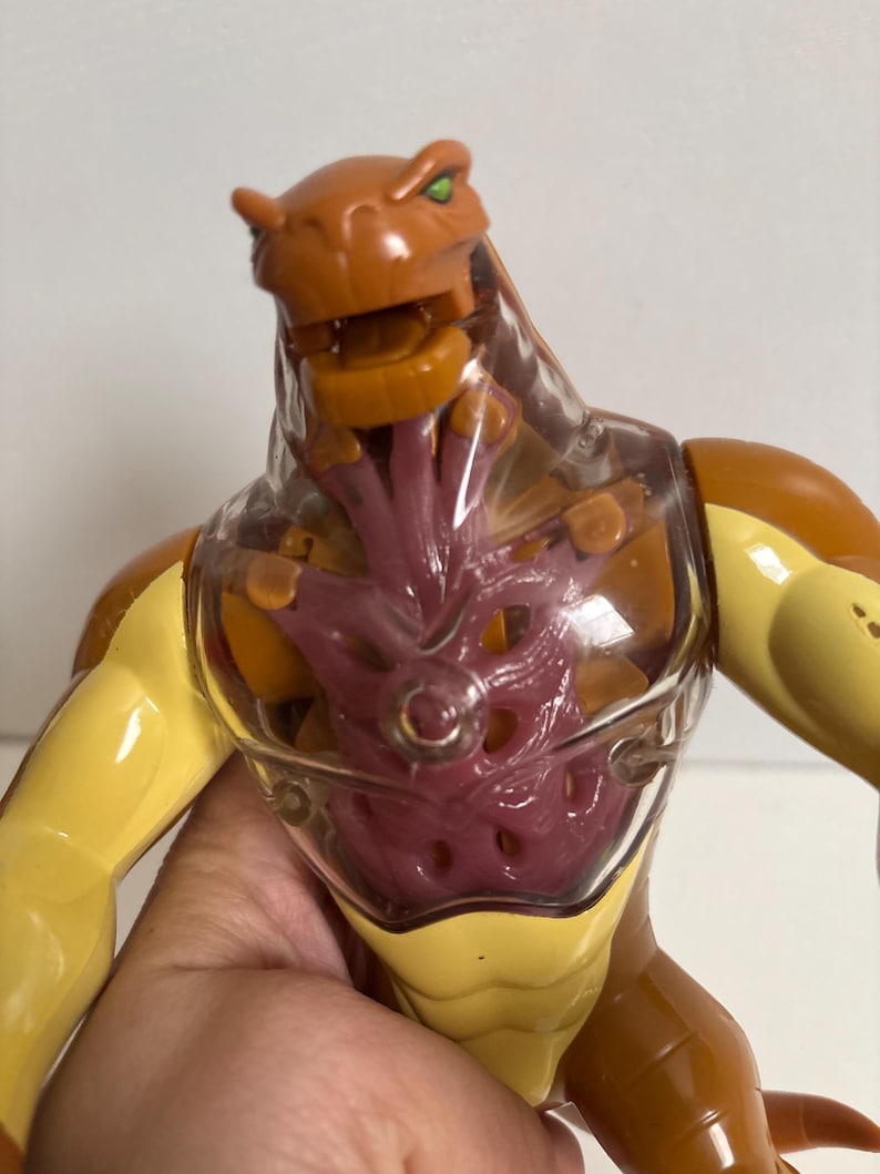 May include: A brown and yellow action figure with a clear chest cavity revealing a purple interior. The figure has a reptilian head with green eyes and is posed with its arms extended. The toy is made of plastic.