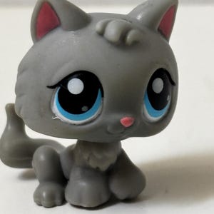 May include: A gray cat figurine with large black eyes, blue irises, and pink inner ears. The cat is in a seated position with its tail curled. The figurine has a small pink nose and a fluffy chest.