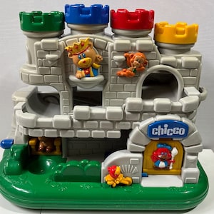 May include: A plastic toy castle with a gray stone-textured facade, featuring four colorful turrets in green, blue, red, and yellow. The structure includes molded animal figures, a yellow door with the blue Chicco logo, and a green base.