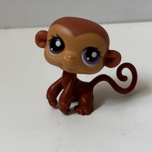 May include: A brown and tan monkey figurine with large, expressive eyes and a curled tail. The toy monkey has a light brown face and a darker brown body, arms, and legs. The monkey is a collectible toy.