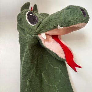 May include: A green fabric alligator hand puppet with white teeth, a red tongue, and black eyes. The puppet has a textured, scale-like pattern and is designed to fit over the hand for play or performance. The interior of the mouth is pink.