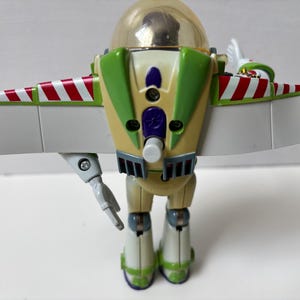 以下が含まれることがあります： 透明なヘルメット、緑と白のアクセント、赤と白のストライプの翼が特徴のバズ・ライトイヤーのアクションフィギュア。おもちゃは可動式の腕と脚を持ち、タンと緑のボディをしています。翼は広げられており、フィギュアは直立しています。