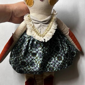 Puede incluir: Mu&ntilde;eca hecha a mano con cabeza de zorro, con un tocado naranja y rojo y detalles circulares amarillos. La mu&ntilde;eca lleva una falda estampada en tonos azules, verdes y morados, con botas rojas y un collar dorado.