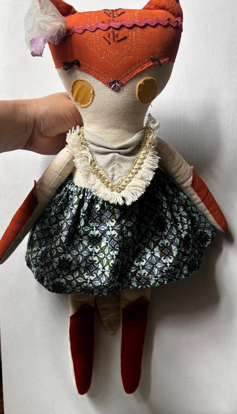 Puede incluir: Mu&ntilde;eca hecha a mano con cabeza de zorro, con un tocado naranja y rojo y detalles circulares amarillos. La mu&ntilde;eca lleva una falda estampada en tonos azules, verdes y morados, con botas rojas y un collar dorado.