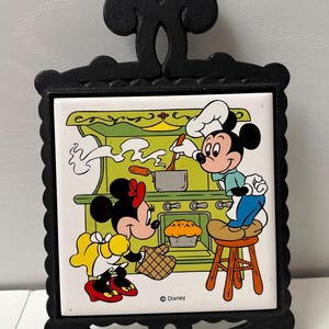 May include: A black cast iron trivet featuring a square ceramic tile insert with a colorful illustration of Mickey and Minnie Mouse cooking in a kitchen. The scene depicts Mickey standing on a stool at a stove while Minnie holds a baking mitt.
