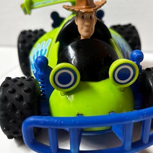 以下が含まれることがあります： ウッディのフィギュアが運転席にいる、緑と青のおもちゃのレーシングカー。車体には大きな黒いタイヤ、青いアクセント、スポイラーが付いています。このおもちゃの車は、コレクターズアイテムです。
