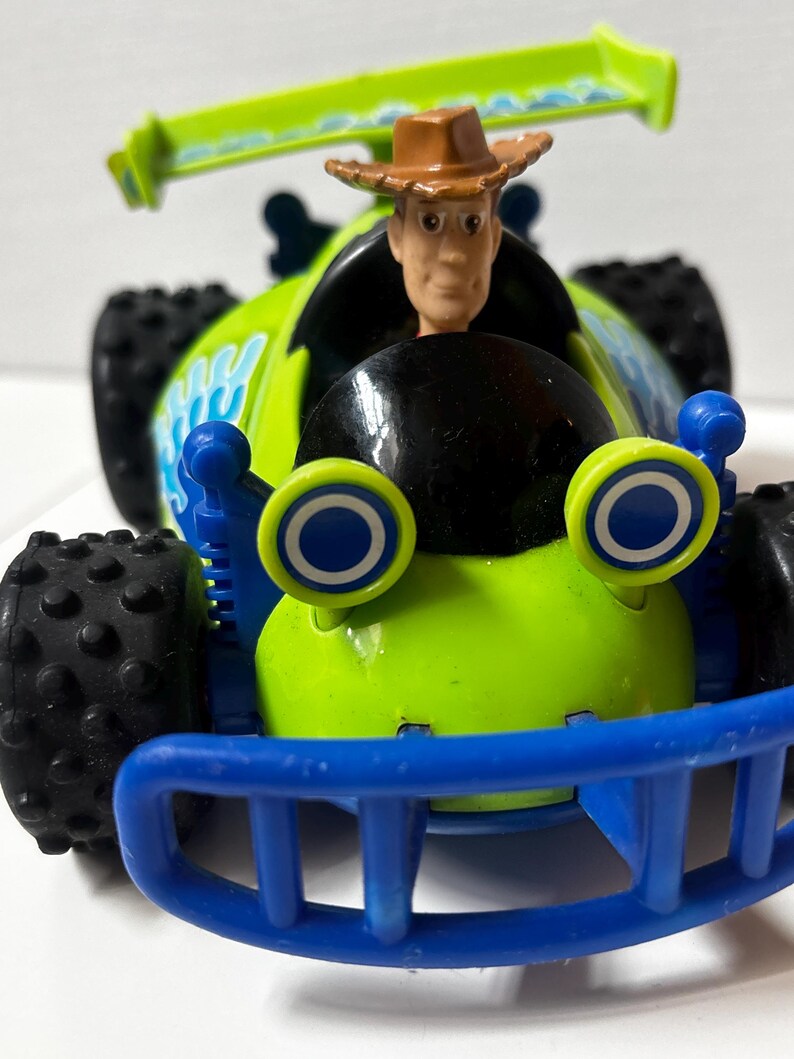以下が含まれることがあります： ウッディのフィギュアが運転席にいる、緑と青のおもちゃのレーシングカー。車体には大きな黒いタイヤ、青いアクセント、スポイラーが付いています。このおもちゃの車は、コレクターズアイテムです。