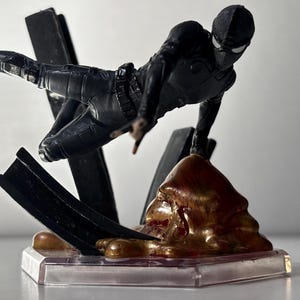 May include: A black action figure of a superhero in a dynamic pose, leaping forward. The figure is on a clear base with a brown and gold textured element and black supports. The superhero is wearing a black suit and mask.