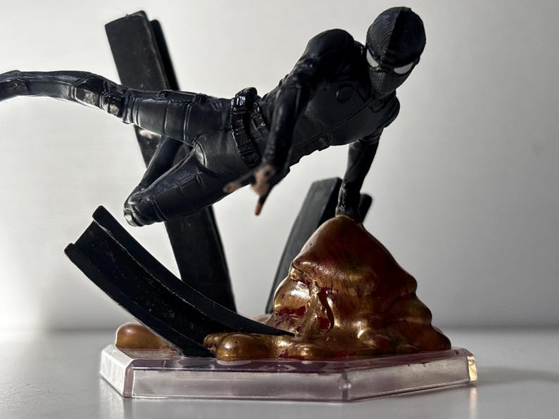 May include: A black action figure of a superhero in a dynamic pose, leaping forward. The figure is on a clear base with a brown and gold textured element and black supports. The superhero is wearing a black suit and mask.