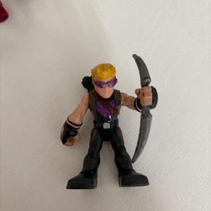 May include: A small plastic action figure of Hawkeye, a superhero character. The figure is wearing a purple and black outfit, with a bow and arrow. The figure has yellow hair and purple goggles.