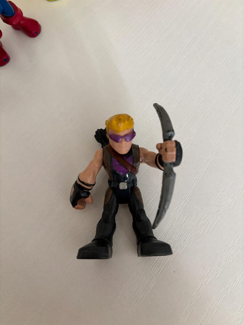 May include: A small plastic action figure of Hawkeye, a superhero character. The figure is wearing a purple and black outfit, with a bow and arrow. The figure has yellow hair and purple goggles.