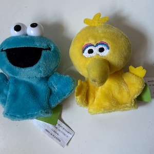 May include: Two plush hand puppets featuring Sesame Street characters. One is a blue monster with large white eyes and a wide black mouth. The other is a yellow bird with a prominent beak, rainbow-colored eyelids, and a small tuft of feathers on top.