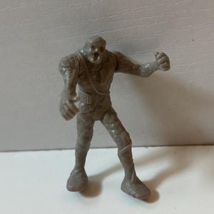 May include: A small, grey figurine of a monster with a textured, stony appearance. The figure has an open mouth, outstretched arms, and detailed leg wrappings. It is a collectible toy or miniature figure.