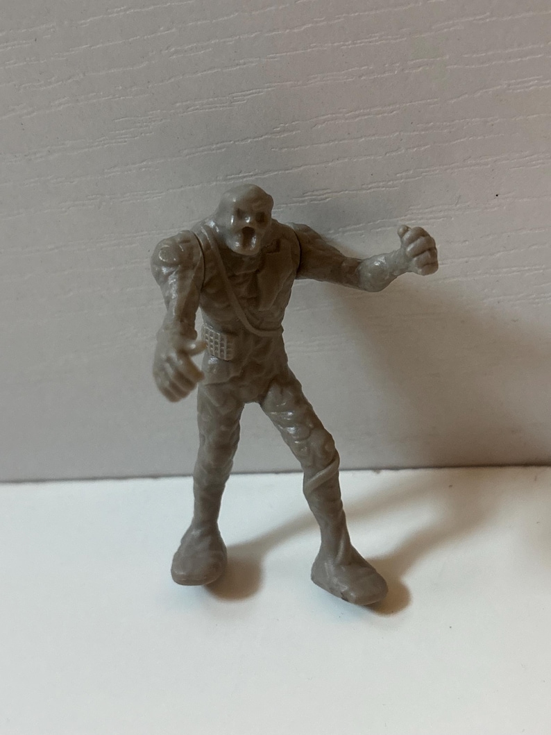 May include: A small, grey figurine of a monster with a textured, stony appearance. The figure has an open mouth, outstretched arms, and detailed leg wrappings. It is a collectible toy or miniature figure.