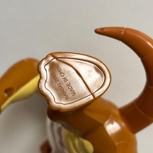 May include: A brown plastic toy figure with a curved tail and a yellow section. The underside of the figure has the text "BANDAI 2008 MADE IN CHINA". The toy is made of shiny plastic.